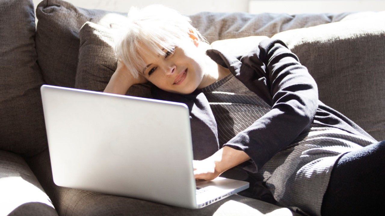A woman lays on a couch smiling at her laptop. 