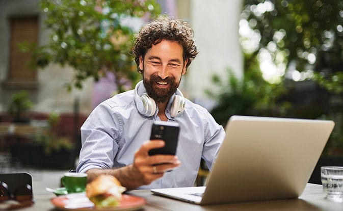 Smiling man holding a smartphone while sitting outdoors with a laptop.