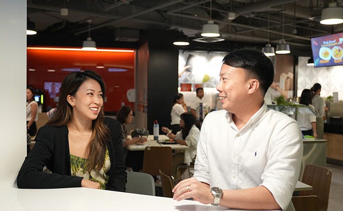 Two coworkers smiling and talking at a table in a casual office setting.