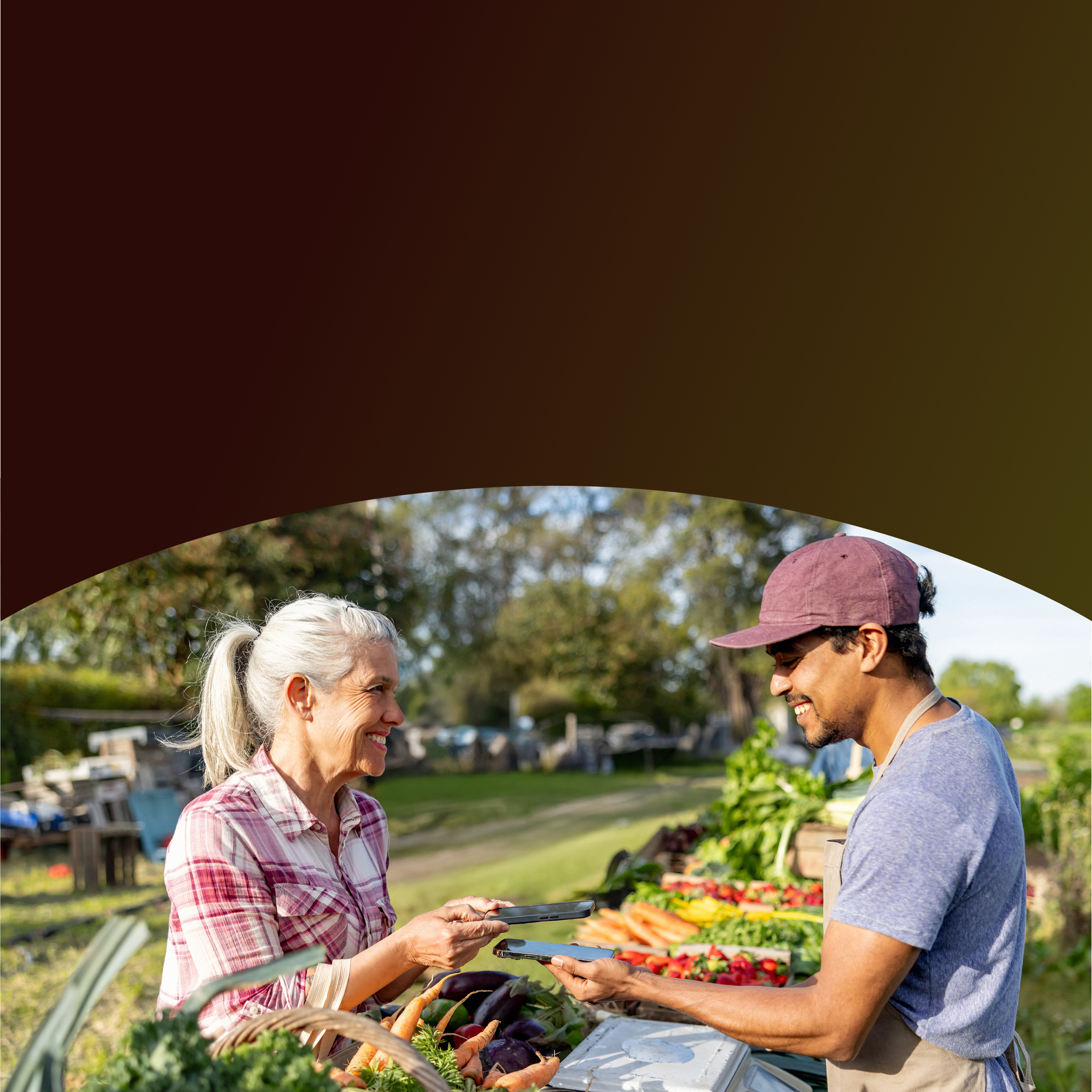 Una cliente sonriente usa su teléfono inteligente para realizar un pago móvil sin contacto a un vendedor que sostiene su teléfono en un puesto de mercado de agricultores al aire libre lleno de productos frescos.