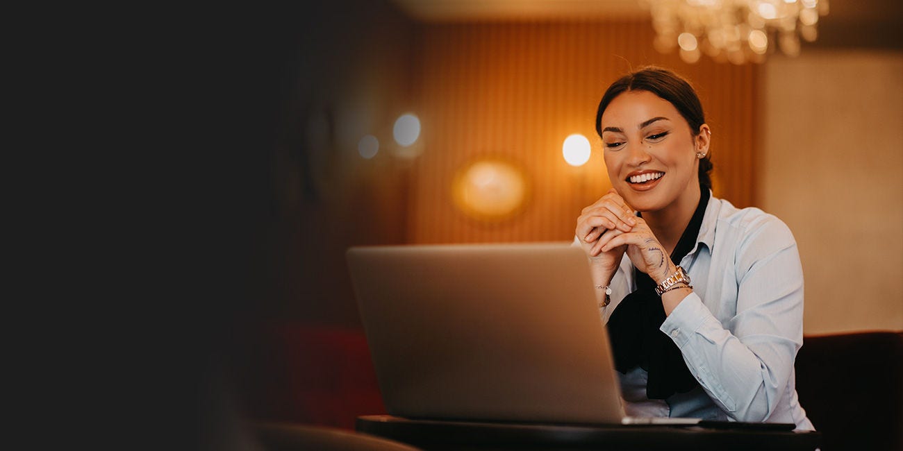 Woman sitting at a table smiling and looking at a laptop in a warmly lit indoor setting.