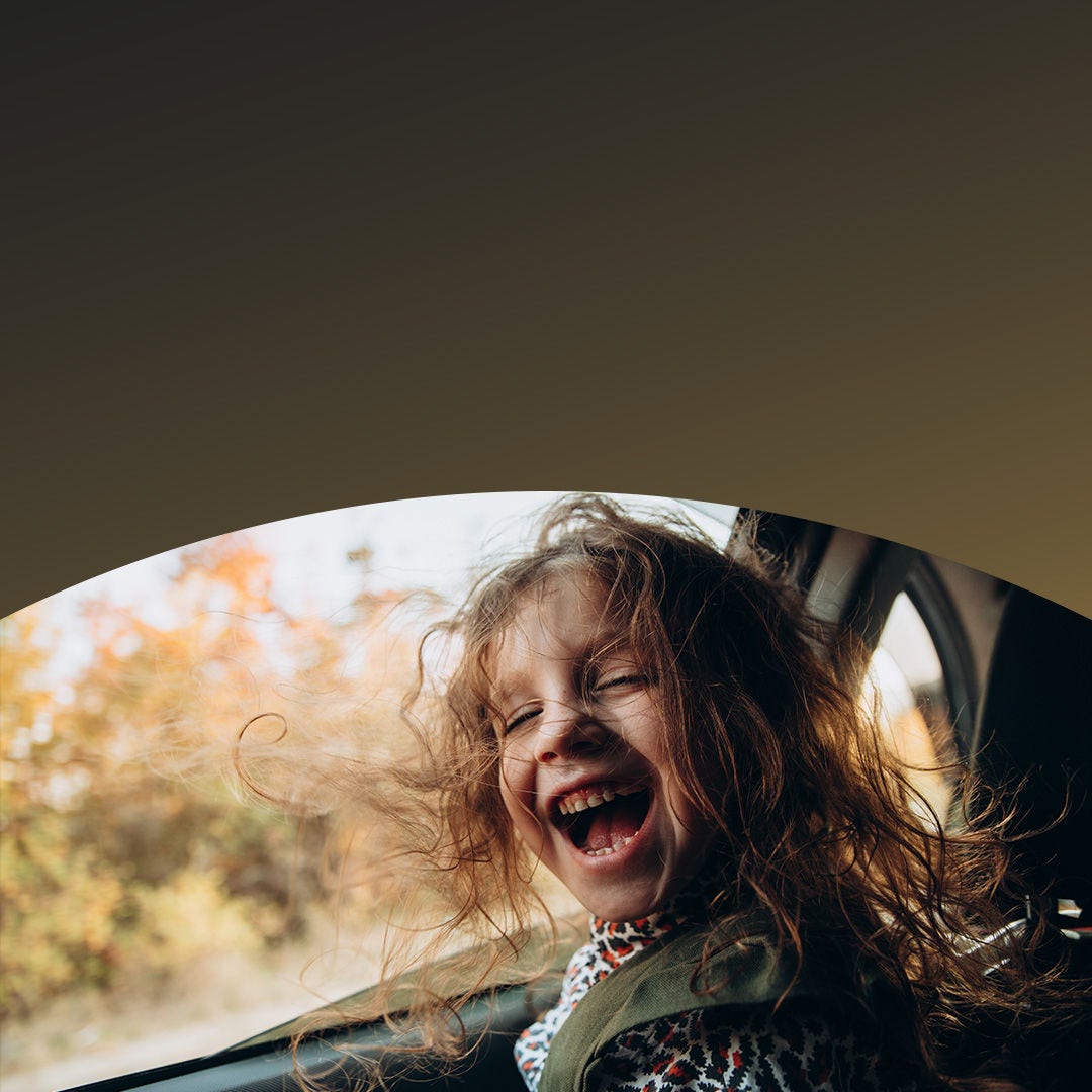Enfant aux cheveux longs riant joyeusement sur la banquette arrière d’une voiture.