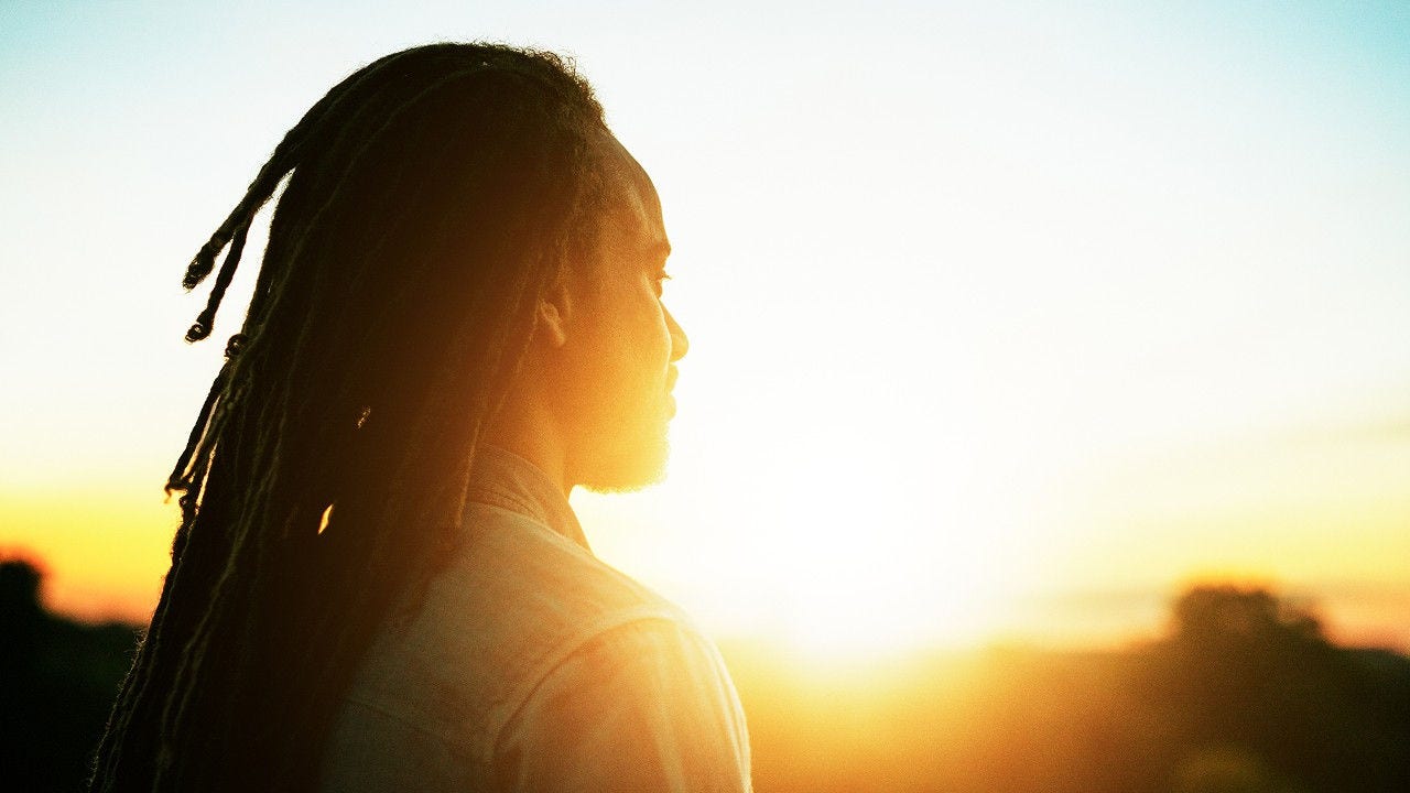 A person with dreadlocks gazes thoughtfully at the sun, surrounded by a serene outdoor setting
