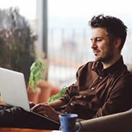 man in brown shirt looking at laptop while having coffee.
