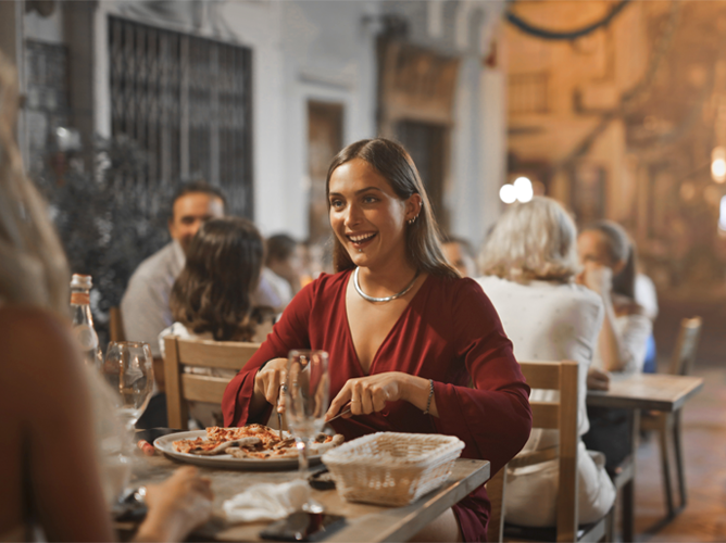 Jeune femme souriante vêtue d'une robe rouge prenant son dîner dans un restaurant animé en plein air, avec des amis en arrière-plan.