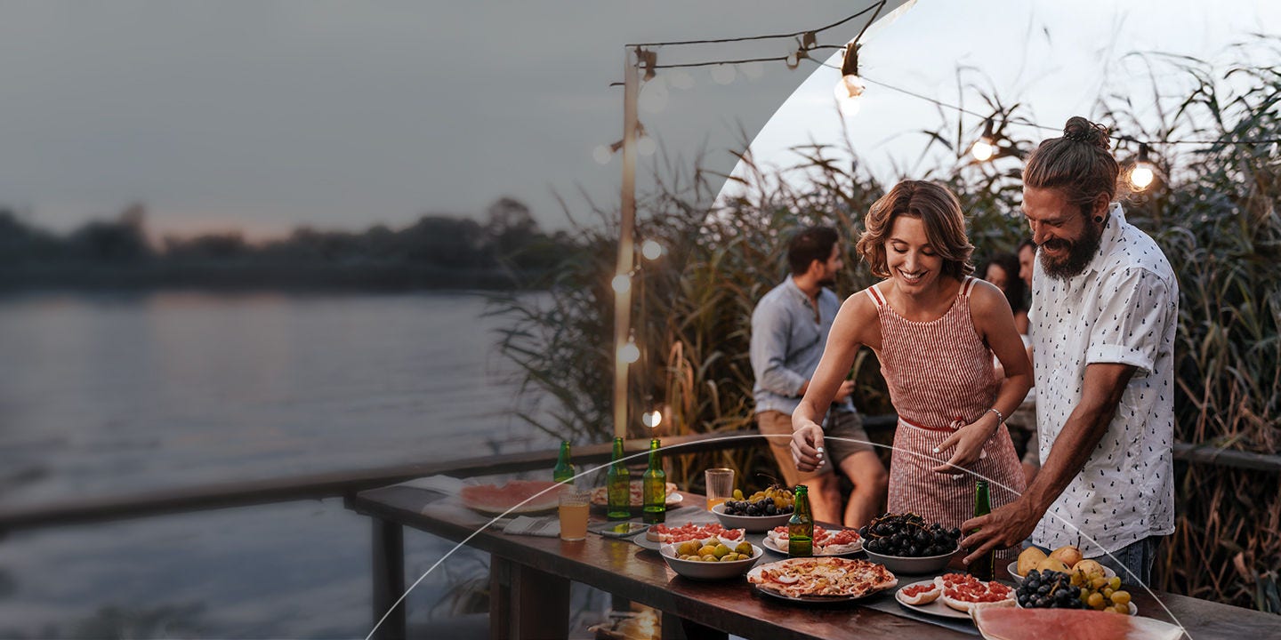 Una pareja sonriente junto a una mesa al lado del lago, con comida, disfrutando de una alegre reunión con amigos.
