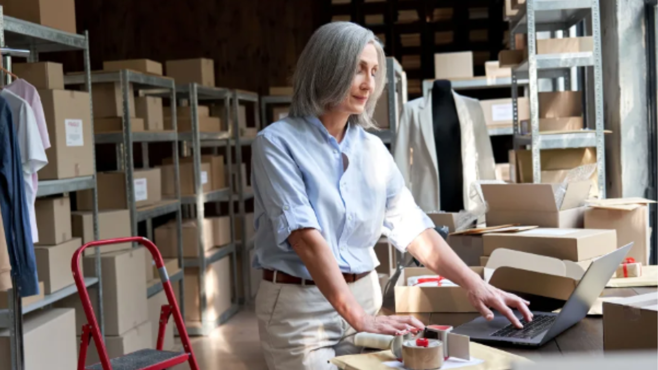 A woman looks at her laptop in a room filled with shelving and boxes. 