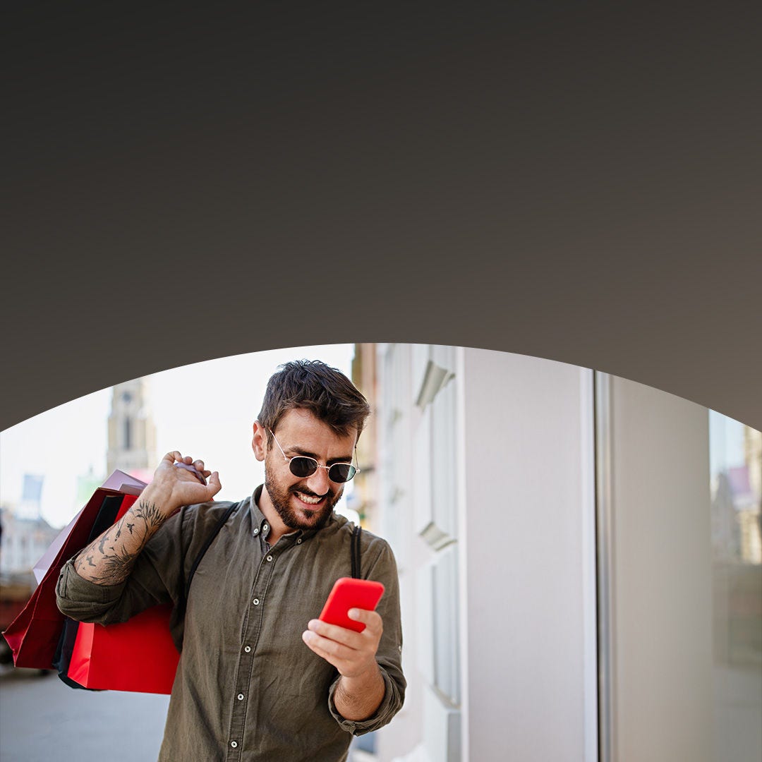 Un homme souriant utilise son smartphone, tenant des sacs de courses dans son autre main.