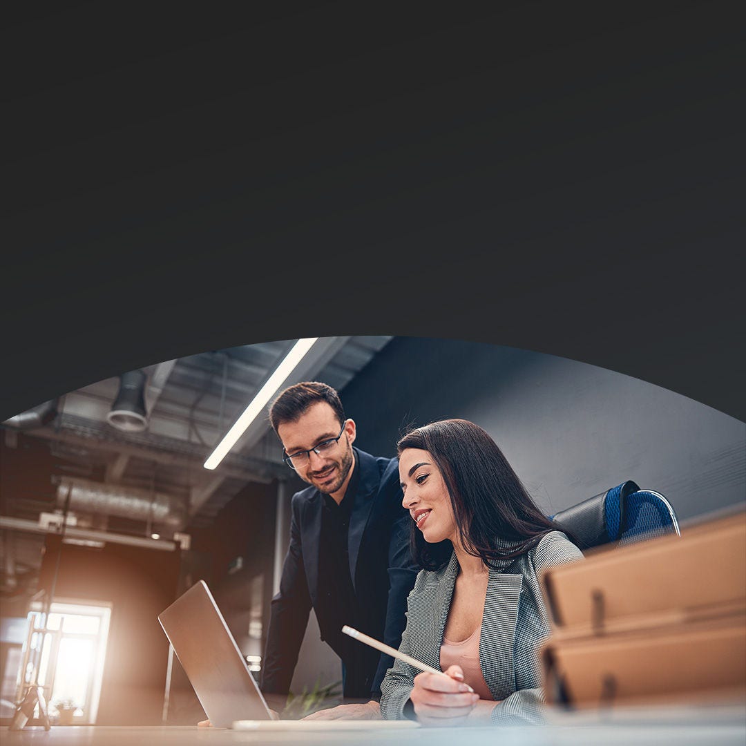 Una mujer sentada en una mesa muestra datos en su computadora portátil a un colega masculino de pie mientras discuten.