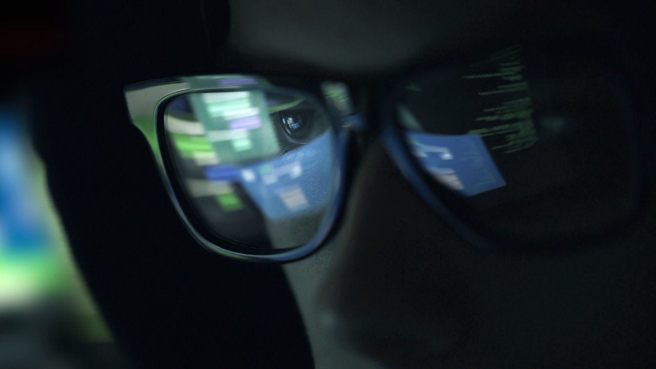 A man in a dark room with computer coding reflected in his eyeglasses. 
