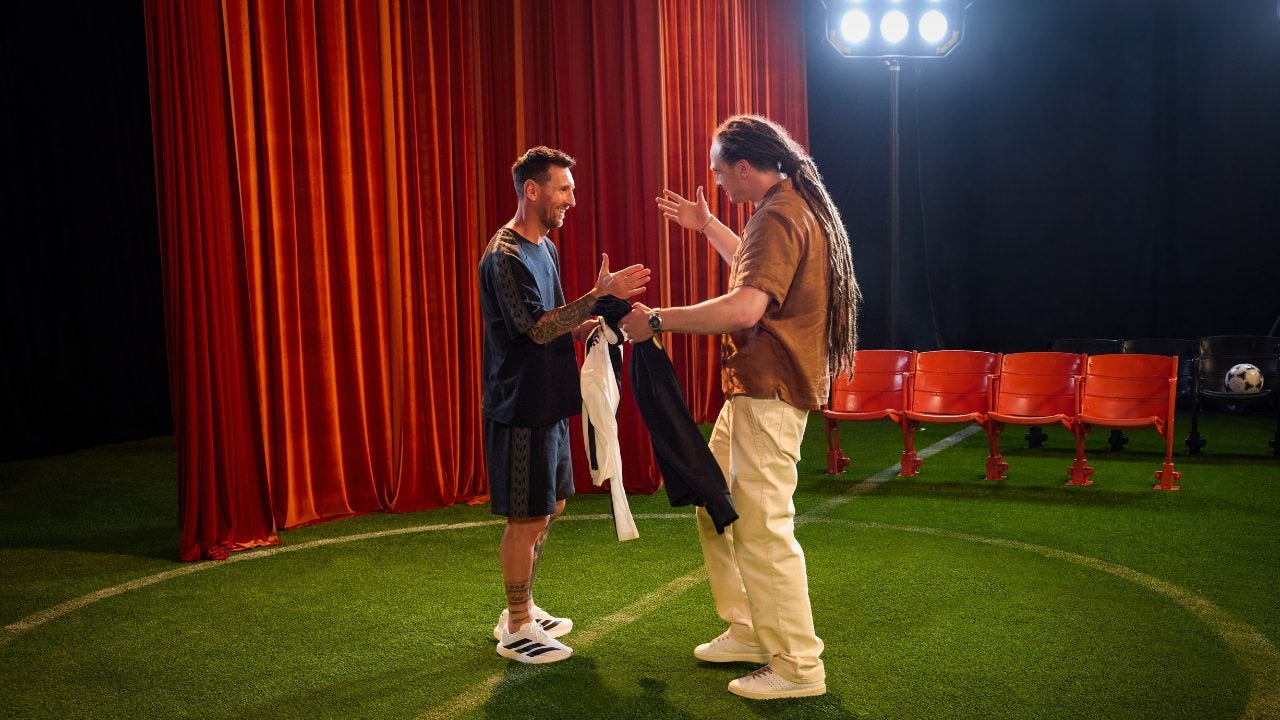 Wyatt Marshall and Lionel Messi are about to shake hands while they hold their jerseys that they are going to swap on a soundstage with stadium seats in the background and on a mock foodtball field.