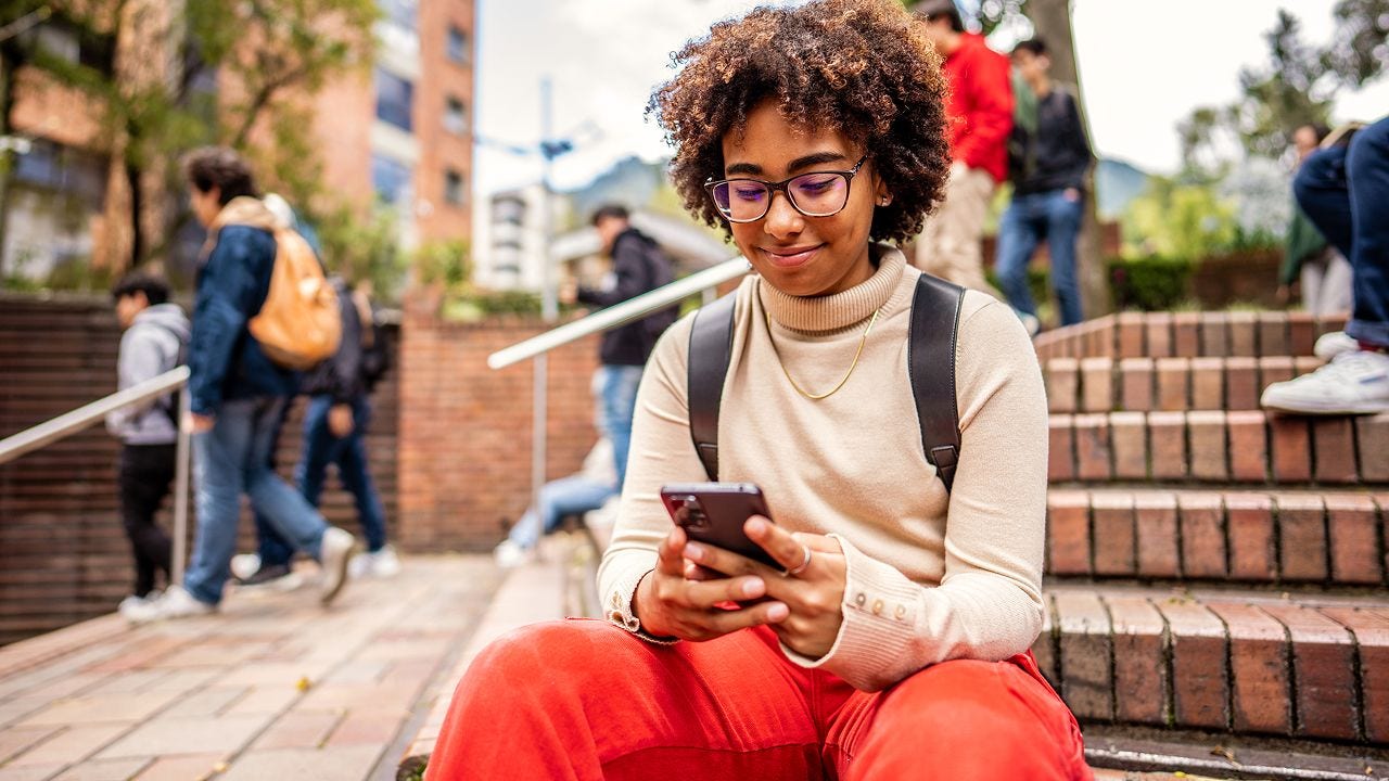A person with curly hair and glasses sits on outdoor steps, looking at their phone and smiling, with other people walking in the background