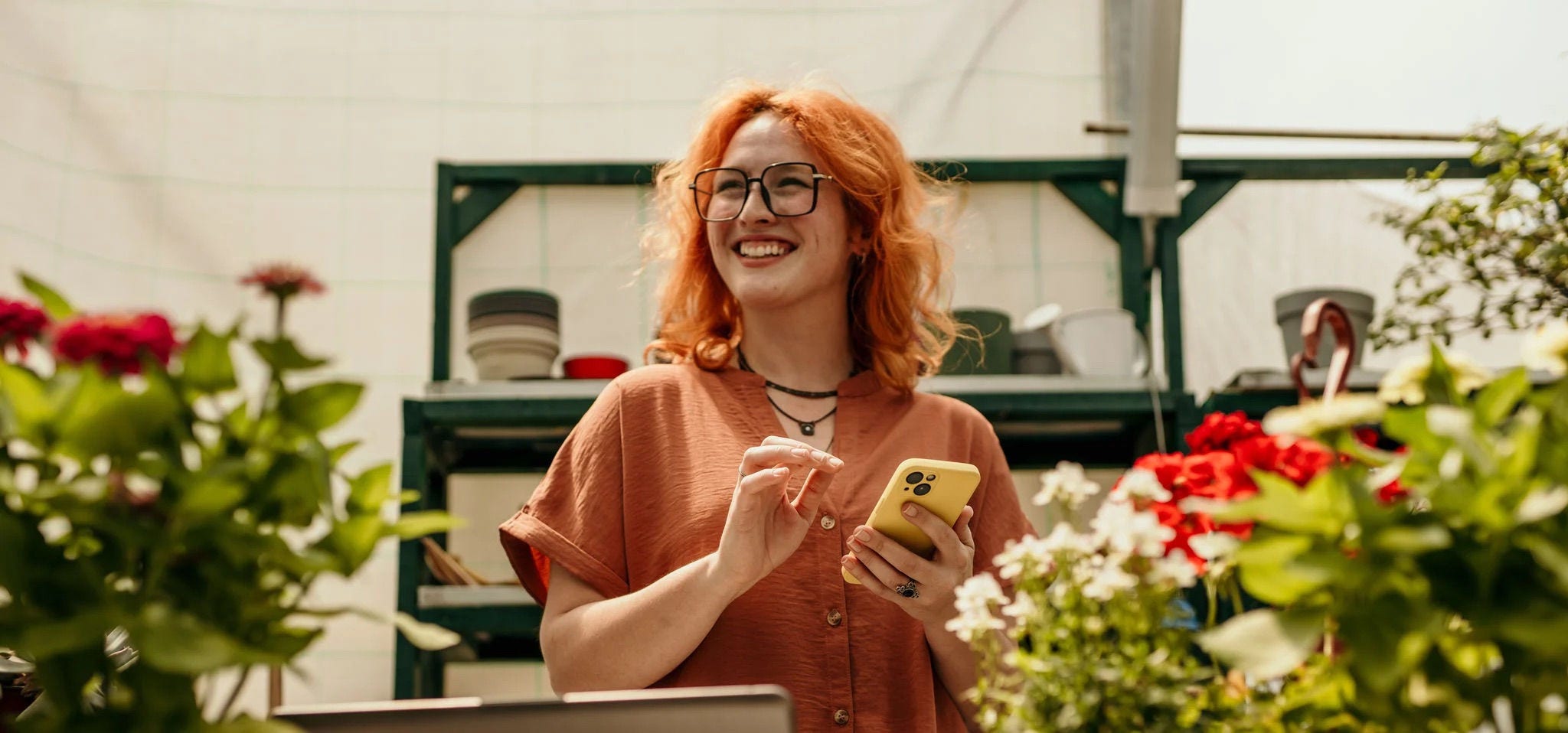 Femme souriante portant des lunettes et utilisant un smartphone dans une pépinière, représentant la propriétaire d’une petite entreprise gérant le commerce électronique ou les interactions avec les clients.