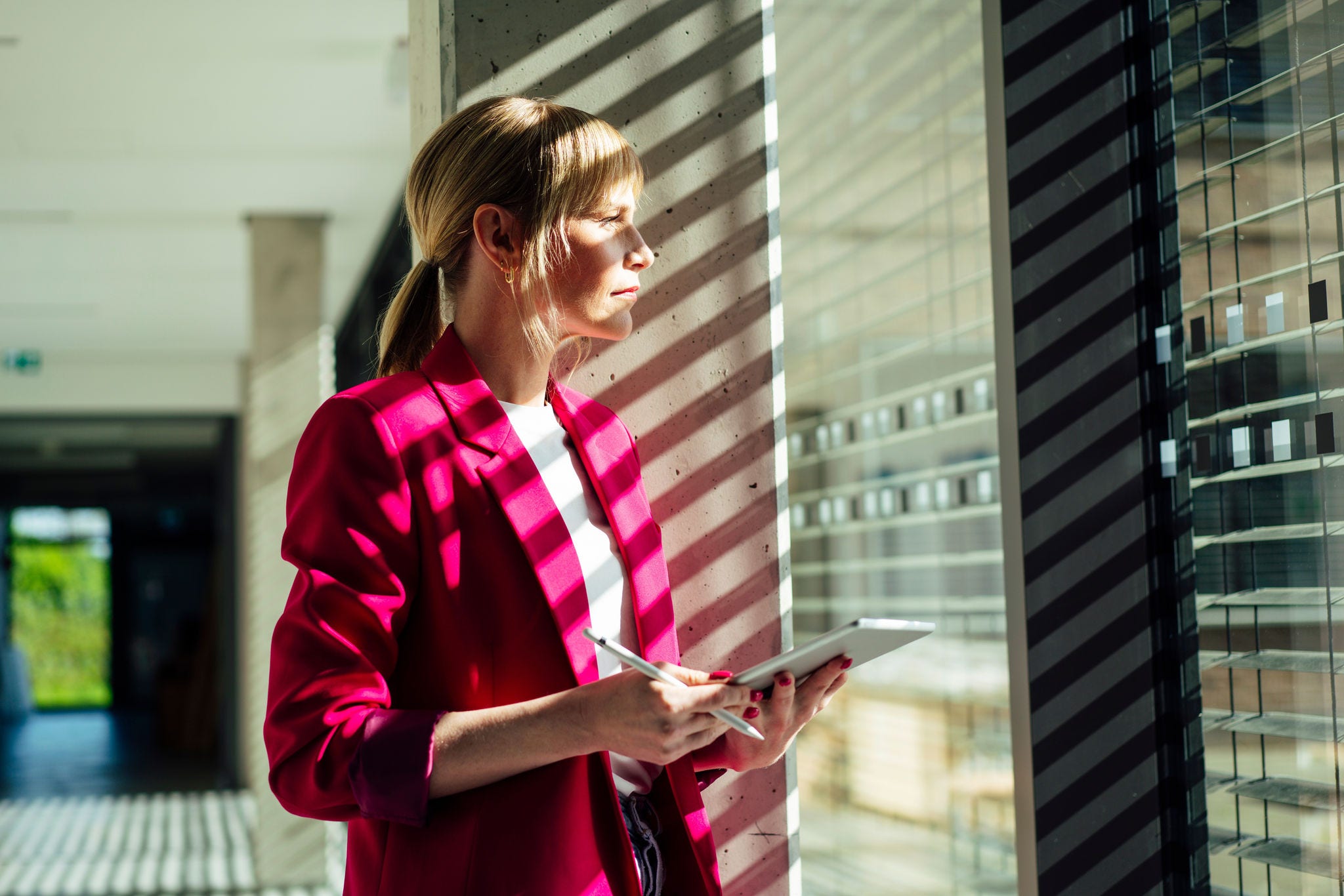 Thoughtful blond businesswoman with tablet pc standing by window at office