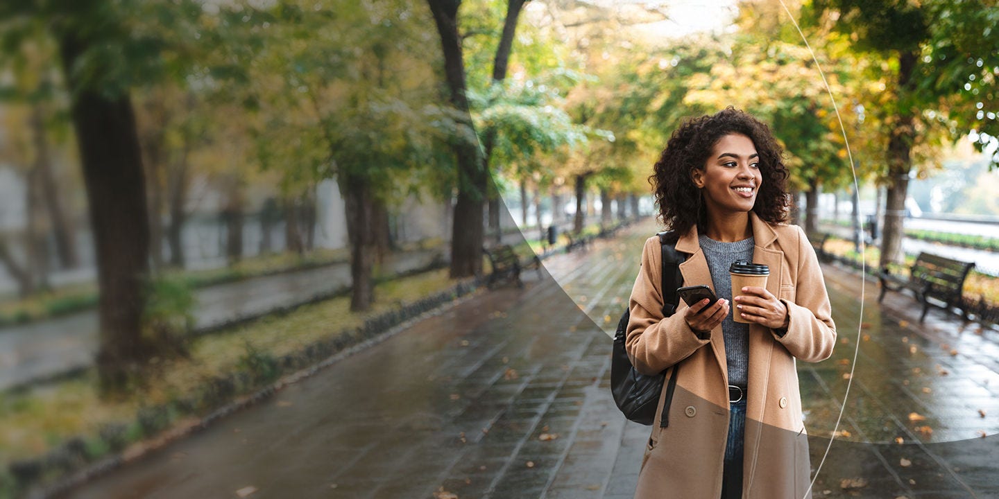 Woman holding a coffee and smartphone whilst walking in a park.
