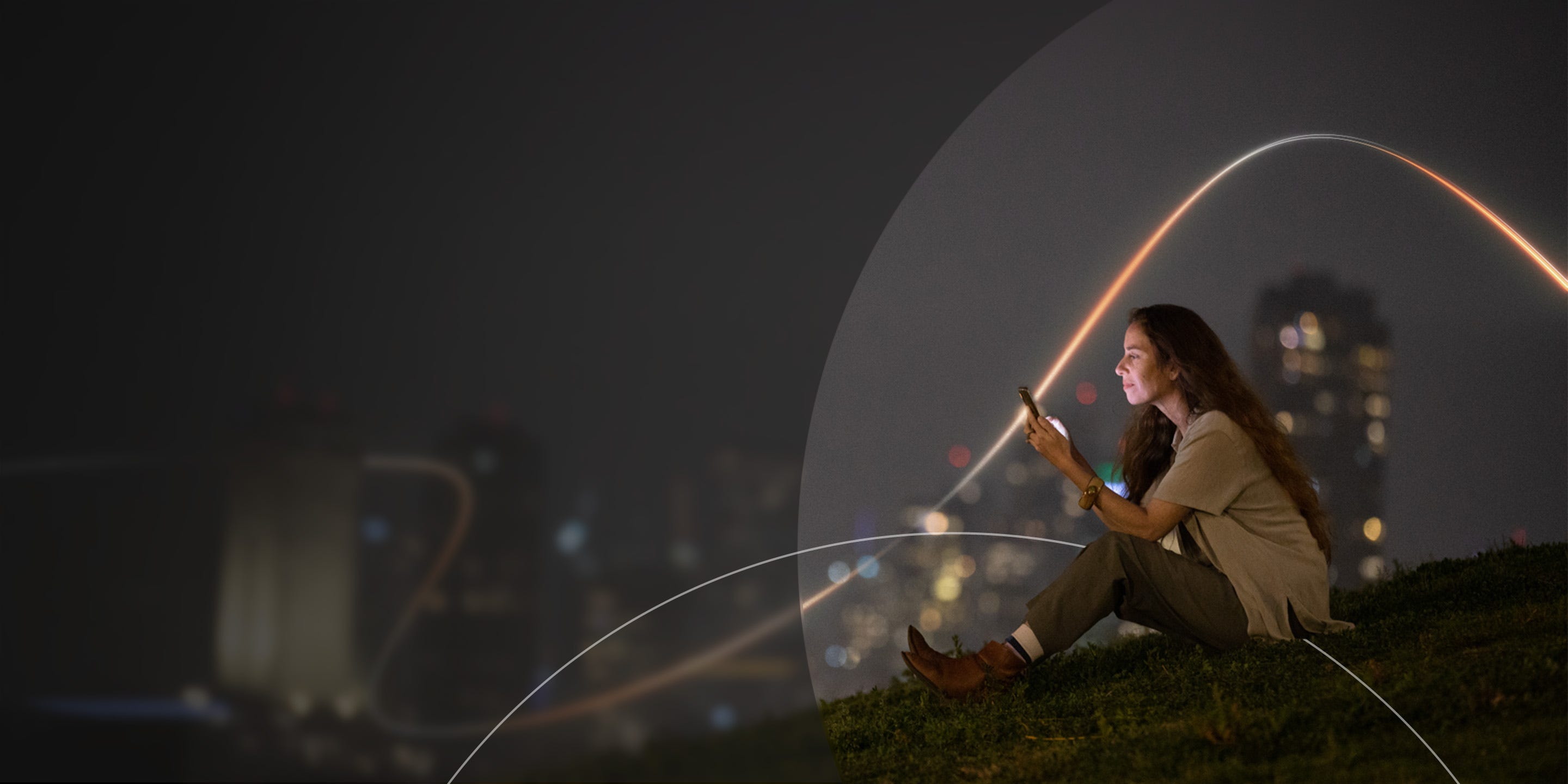 Woman sitting on grass at night, using her phone, with blurred city lights glowing in the background.