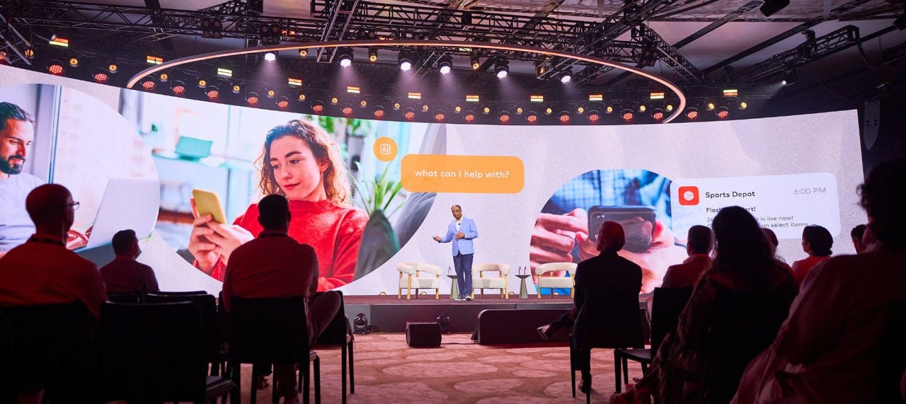 Chiro Aikat speaks on stage in front of a wide image of consumers on their phones at Connections. 