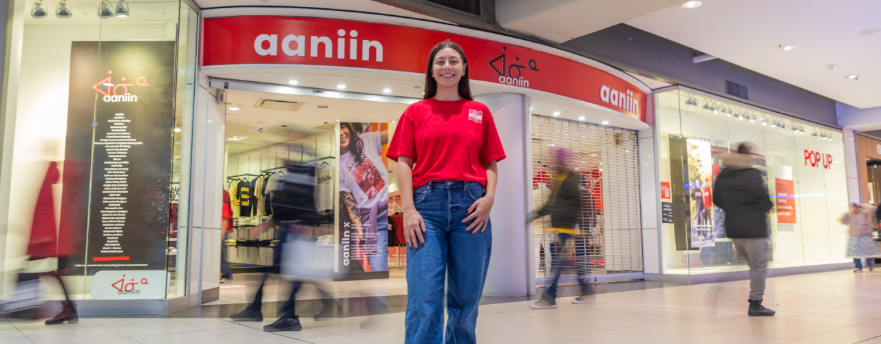 Chelsee Pettit porte une chemise rouge et un jean devant son grand magasin, Aaniin, tandis que les clients marchent derrière elle. 